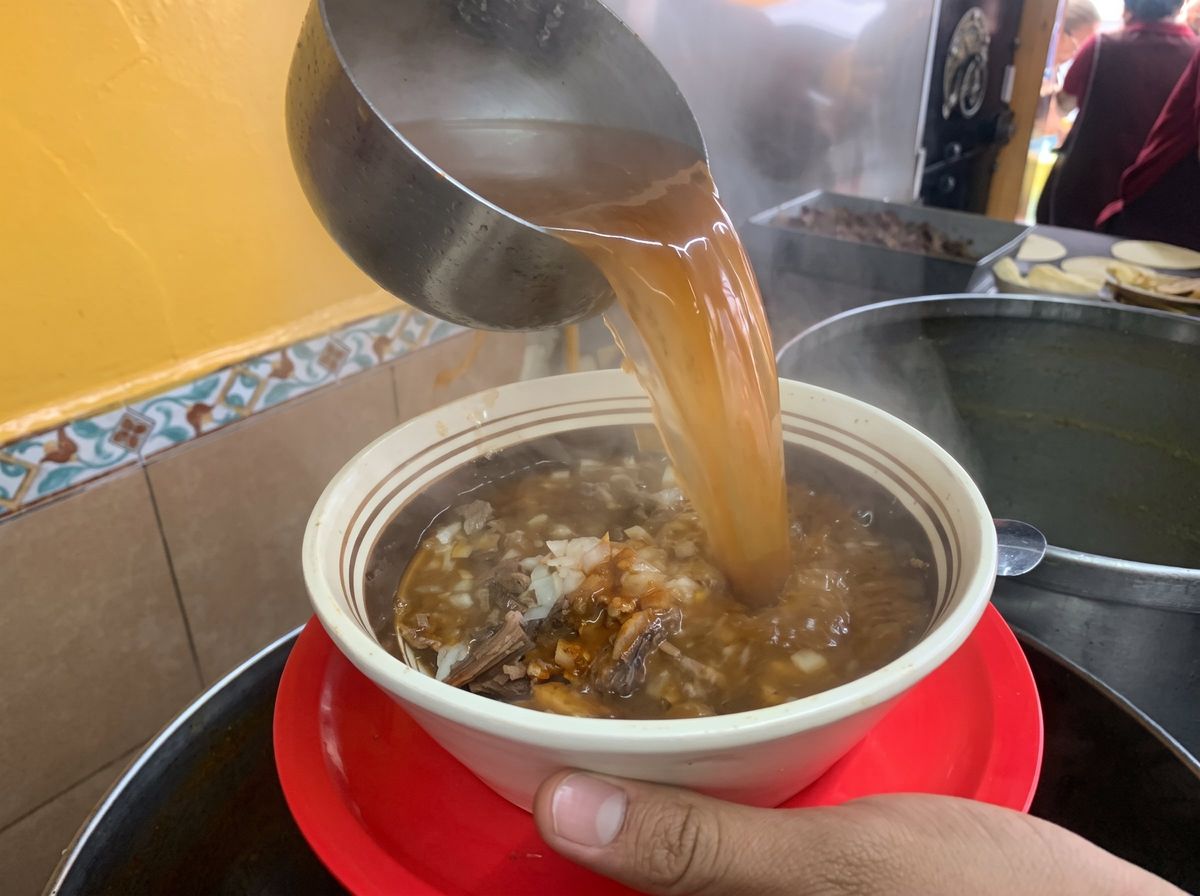 Cucharón vertiendo consomé humeante sobre un tazón de birria en la cocina